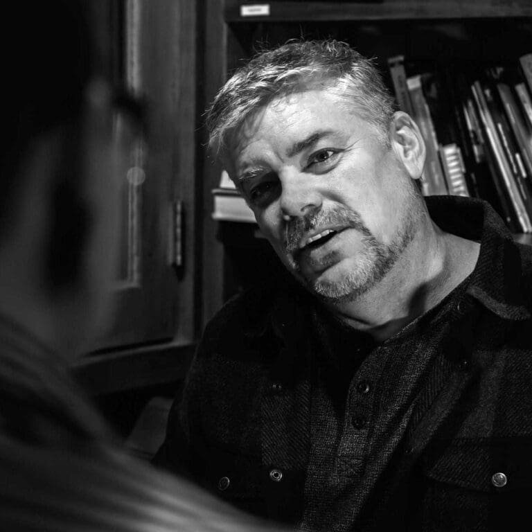 Dan Nachtrab Voice Actor sitting in front of wall of books and speaking with person (blurred out) across a table